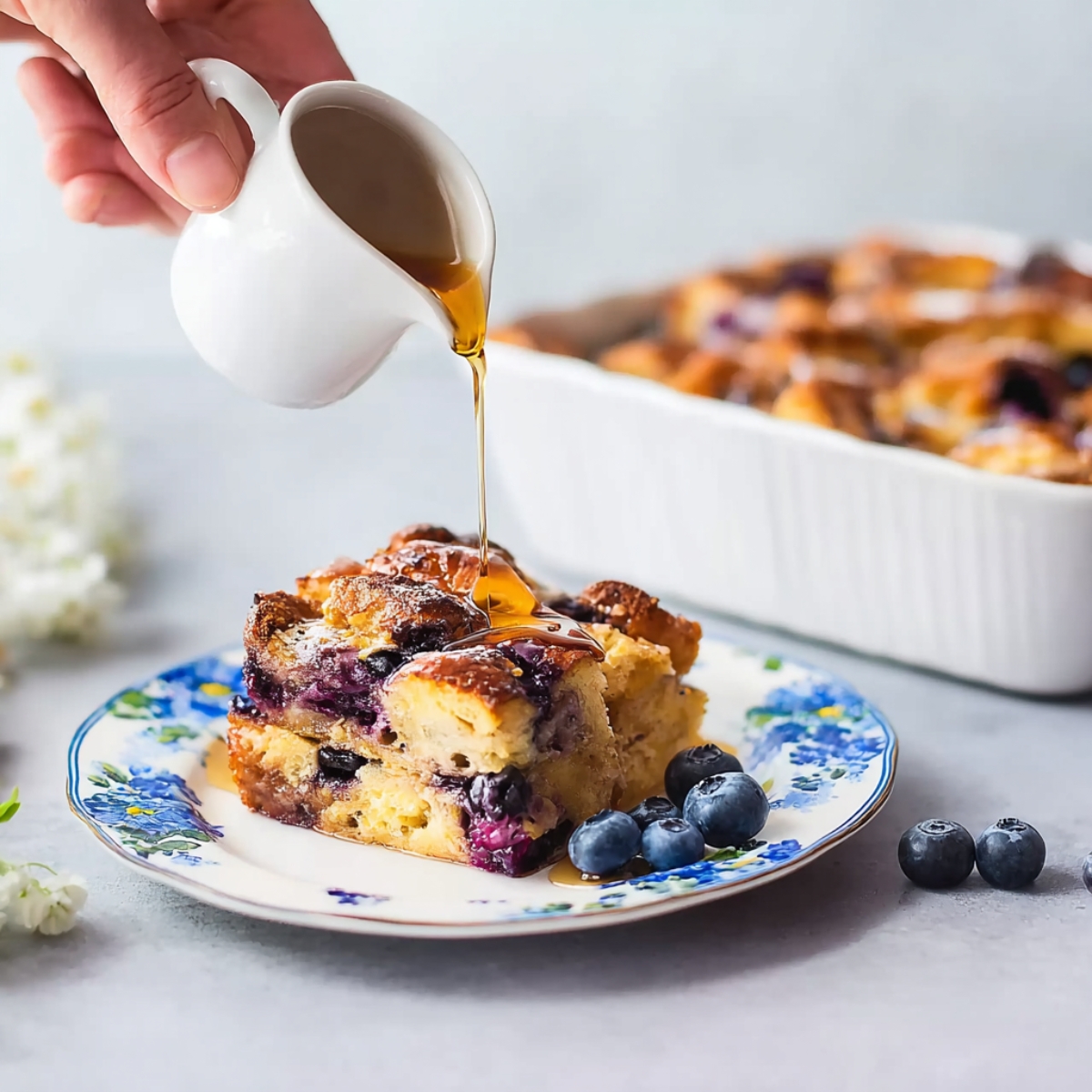 Slice of blueberry French toast casserole drizzled with warm maple syrup on a floral plate, with fresh blueberries on the side.
