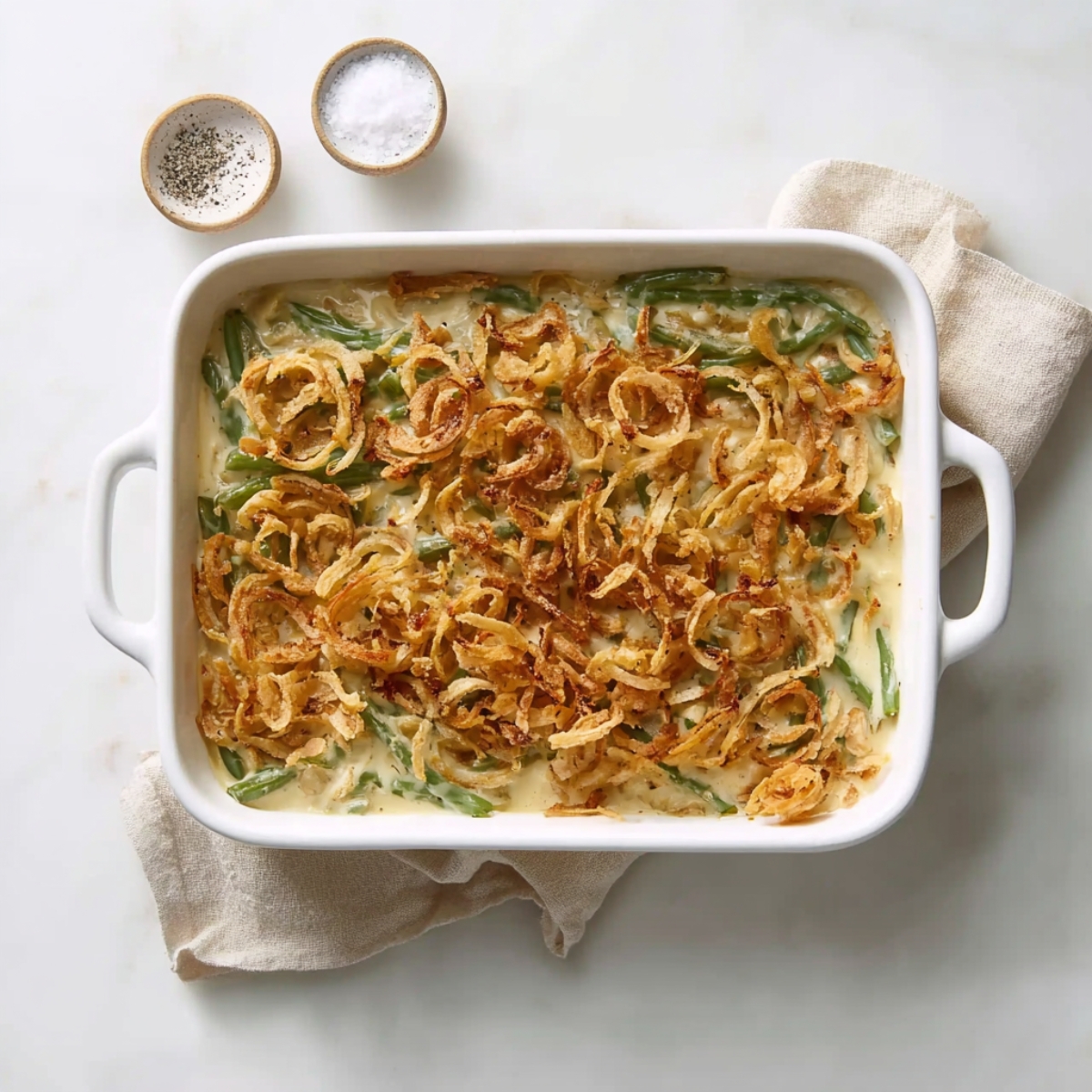 Overhead view of creamy homemade green bean casserole topped with crispy fried onions in a white baking dish on a neutral background