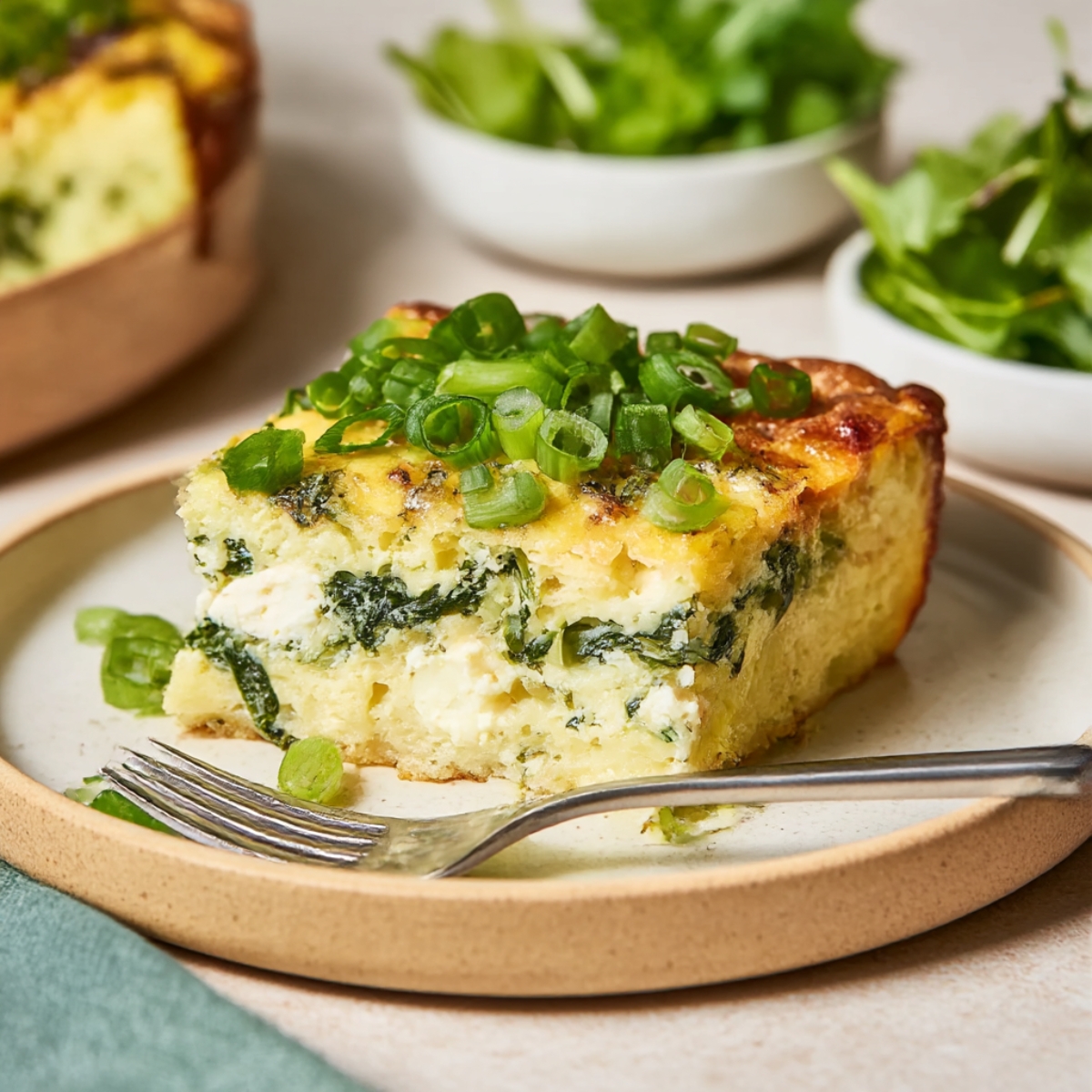 A slice of baked cottage cheese egg bake, topped with fresh green onions, served on a plate with a fork, alongside a side of mixed salad greens.