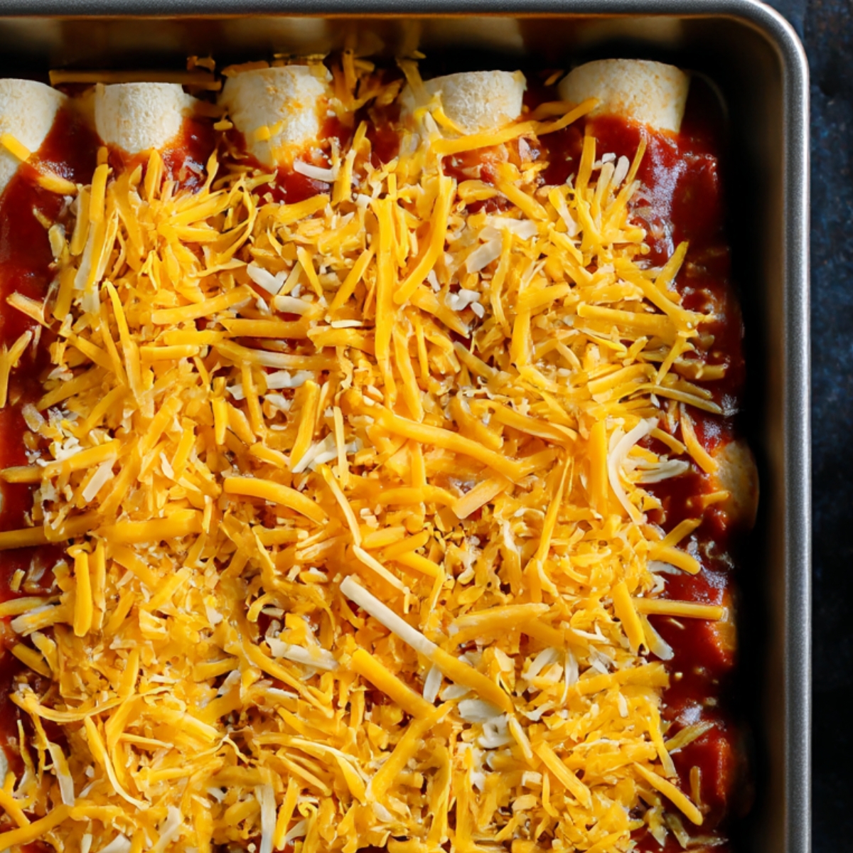 A close-up of chicken enchiladas topped with a blend of shredded cheddar and mozzarella cheese, covered in red sauce, ready to be baked until golden and bubbly.