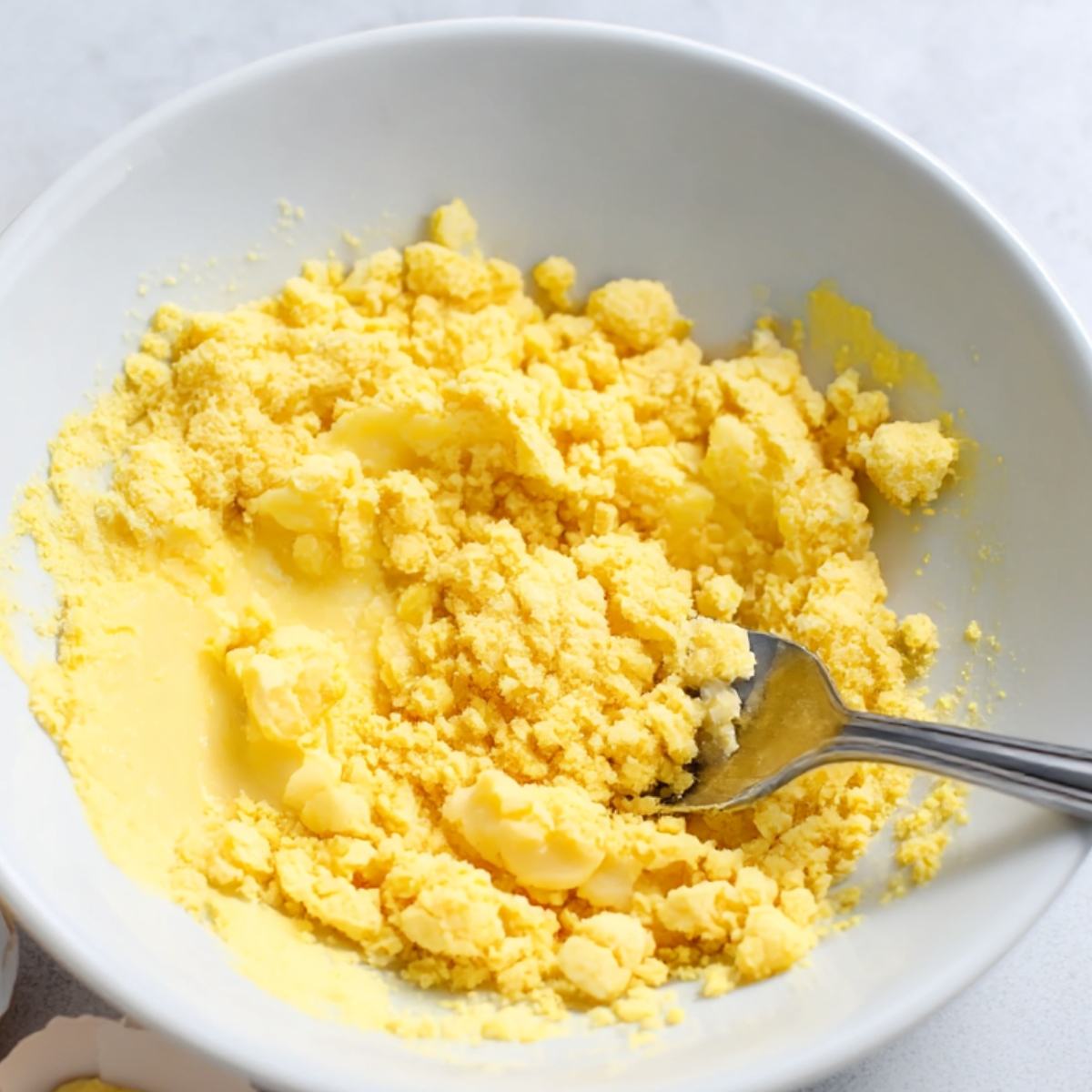 A close-up of crumbled egg yolk in a white bowl, prepared for deviled egg pasta salad, showing the texture of the yolk being mashed and ready to be mixed into the creamy dressing for the salad.