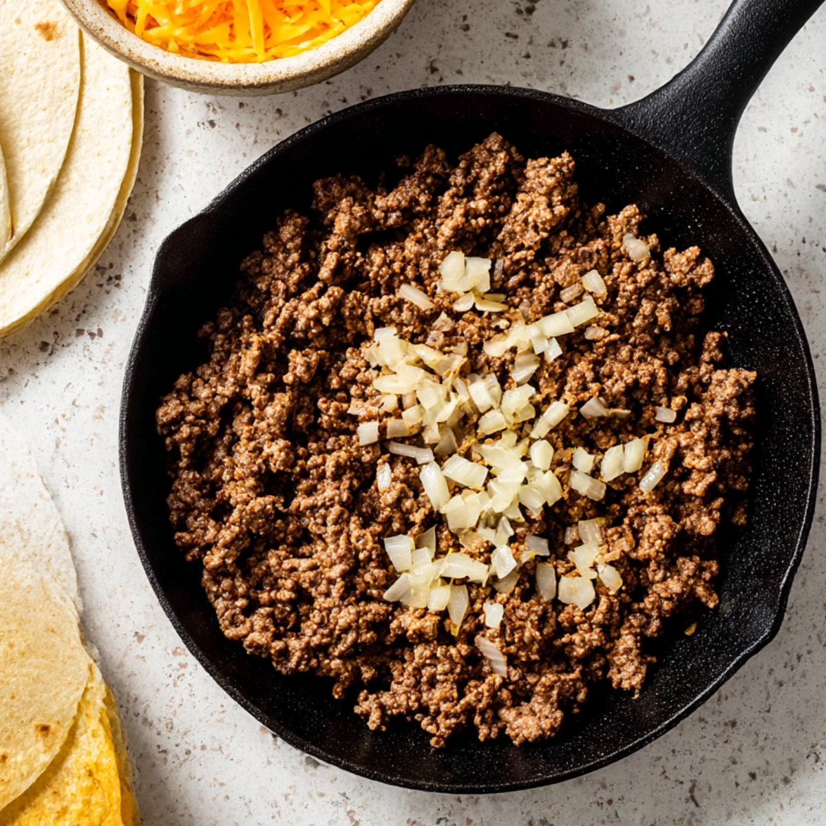 Crumbled ground beef and diced onions cooking together in a cast iron skillet, perfect for taco filling or other ground beef recipes, served with tortillas and cheese.