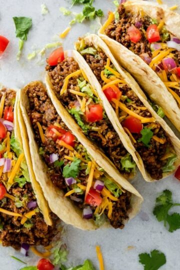 Delicious ground beef soft tacos topped with shredded lettuce, cheddar cheese, diced tomatoes, and onions, served in soft flour tortillas, perfect for a quick and tasty meal.