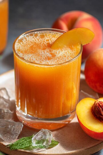 A glass of homemade peach juice with ice cubes and a slice of peach, surrounded by fresh peaches and a sprig of mint on a rustic plate.