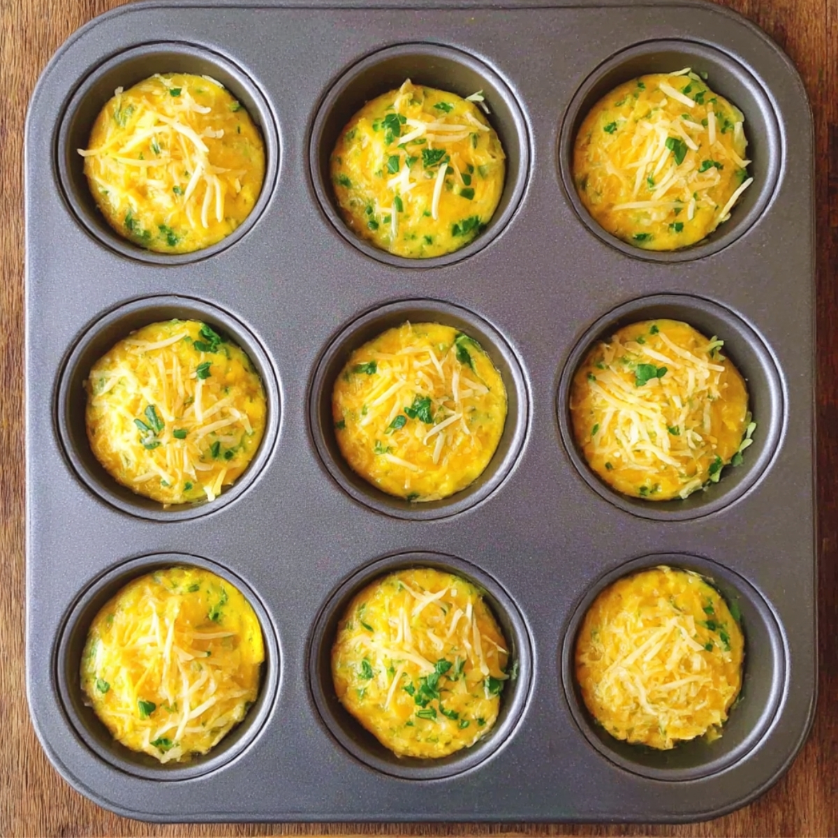 A tray of freshly baked Starbucks-style egg bites, topped with shredded cheese and chopped herbs, golden and puffed up in a muffin tin. These bite-sized treats are ready to enjoy!