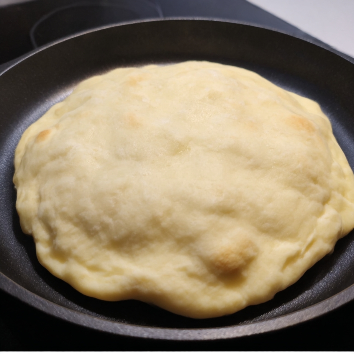 A piece of Turkish bread cooking in a skillet, puffing up as it heats. The dough is rising and starting to form light golden brown spots, showing the early stages of cooking before it is fully browned and ready to serve.