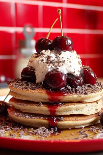 Close-up of Cherry Chocolate Pancakes stacked high and topped with fresh cherries, whipped cream, chocolate shavings, and cherry syrup dripping over fluffy golden pancakes on a red plate.