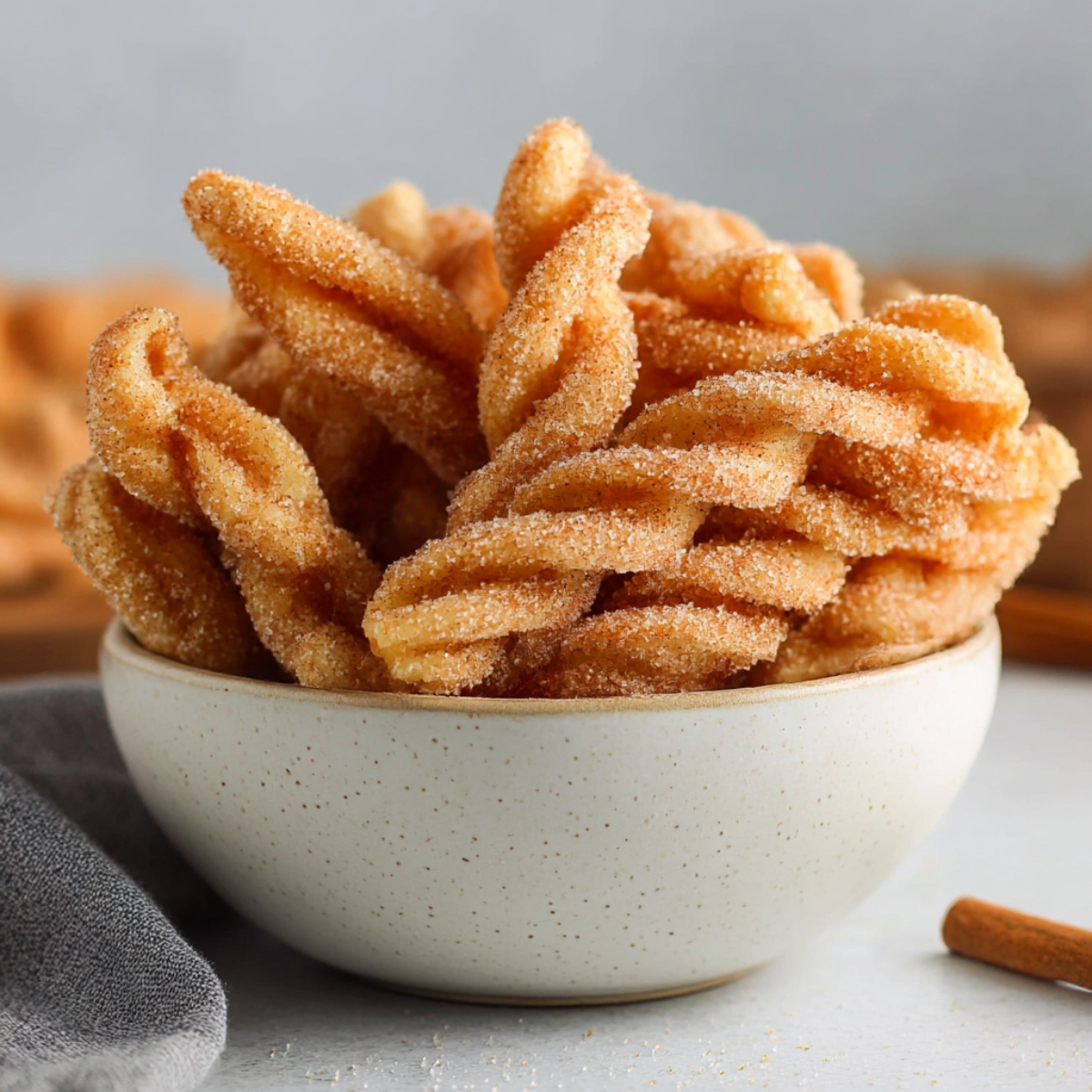 A close-up of golden, crispy cinnamon twists coated in a sweet cinnamon-sugar mixture, served in a white speckled bowl, ready to enjoy.