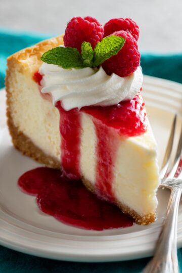 Close-up of a creamy cheesecake recipe slice topped with fresh raspberries, whipped cream, and a generous drizzle of raspberry sauce, served on a white plate.