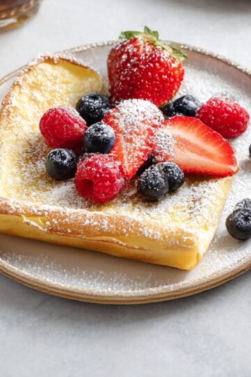 German pancakes (Dutch baby) baked until golden and puffy, topped with fresh strawberries, blueberries, raspberries, and powdered sugar on a ceramic plate.