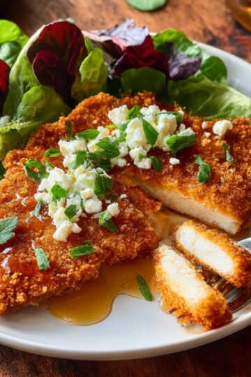 Close-up of crispy honey feta chicken breast with melted feta cheese and honey sauce served alongside fresh salad greens.