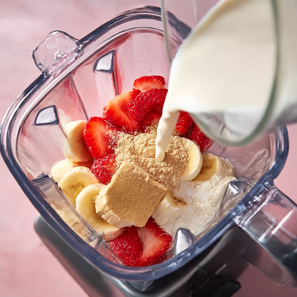 Strawberry cheesecake protein shake ingredients in a blender with sliced strawberries, banana, protein powder, and almond milk being poured in