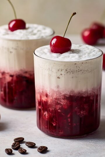 Two glasses of Cherry Vanilla Latte with whipped cream on top, fresh cherries garnish, and a rich cherry syrup layer at the bottom, served on a light background with coffee beans scattered nearby.