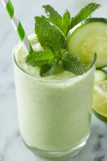 Close-up of a glass filled with creamy cucumber smoothie, garnished with a cucumber slice, fresh mint leaves, and a green-striped straw, with lime halves in the background, perfect for a healthy refreshing drink.