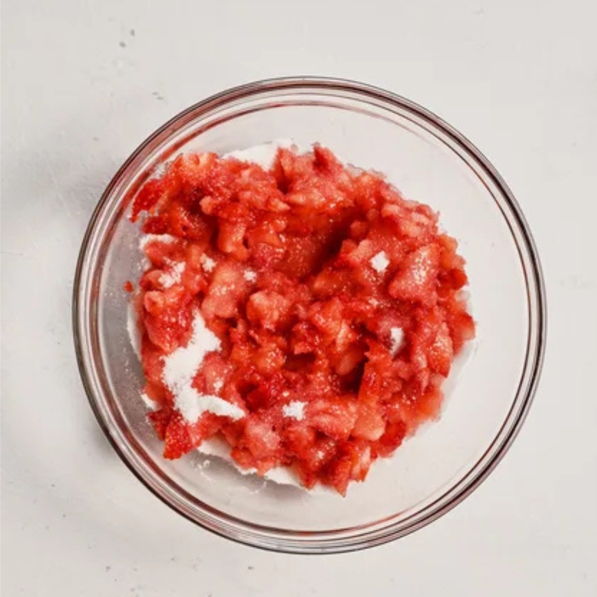Top-down view of a glass bowl filled with chopped strawberries mixed with sugar, forming the sweet base for a festive Strawberry Merry Cream beverage.