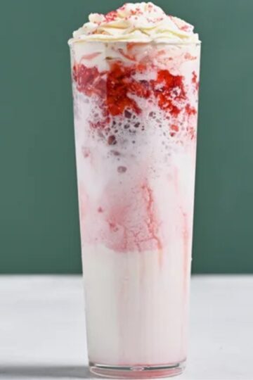 Tall glass of Strawberry Merry Cream layered with pink strawberry cream, whipped cream on top, and sprinkled strawberry bits, served on a light surface with a green background, showcasing a festive creamy strawberry beverage.