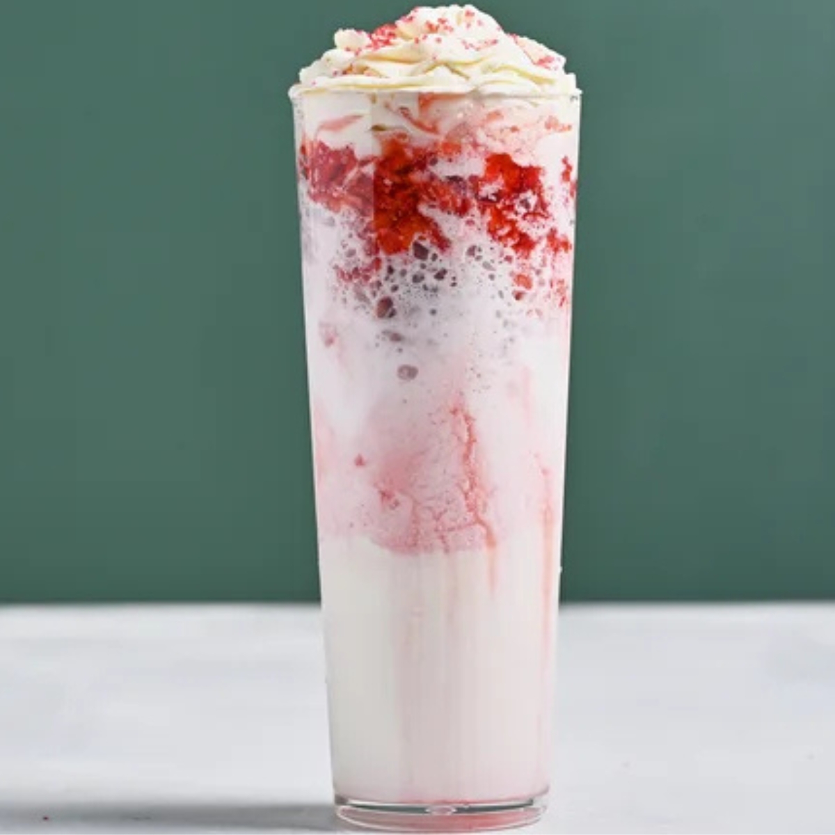 Tall glass of Strawberry Merry Cream layered with pink strawberry cream, whipped cream on top, and sprinkled strawberry bits, served on a light surface with a green background, showcasing a festive creamy strawberry beverage.