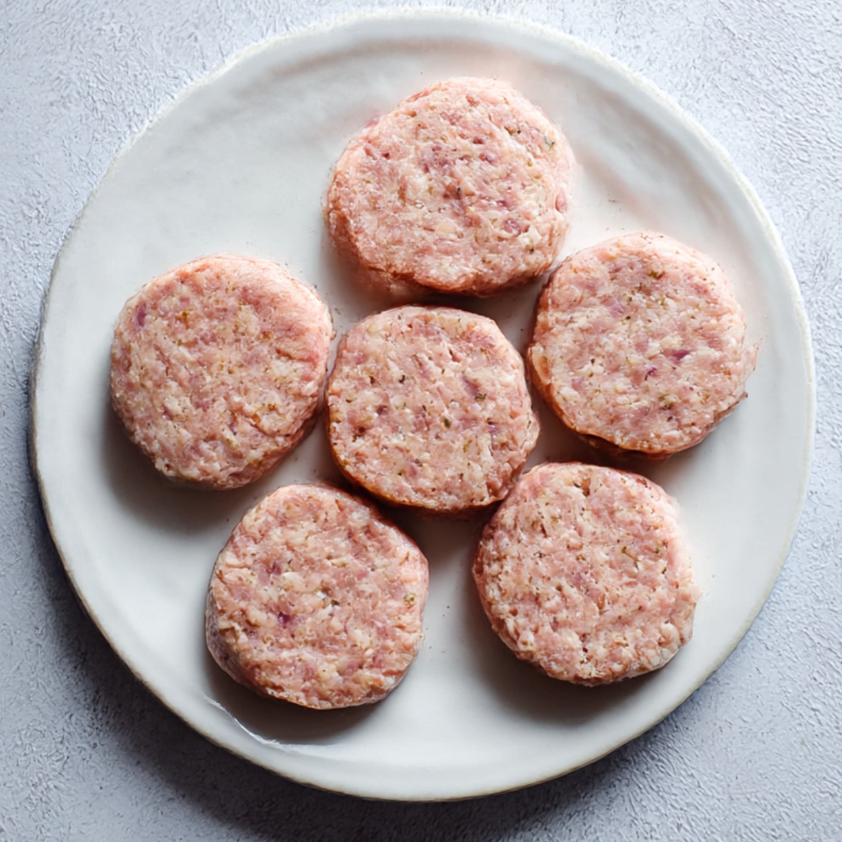 Seven uncooked turkey sausage patties neatly arranged on a white plate, ready for pan-frying or grilling