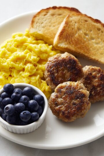 Plate of breakfast featuring golden-brown turkey sausage patties, fluffy scrambled eggs, toasted bread, and a small bowl of fresh blueberries