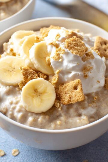 A bowl of creamy banana overnight oats topped with fresh banana slices, whipped cream, and crumbled graham crackers for added crunch.