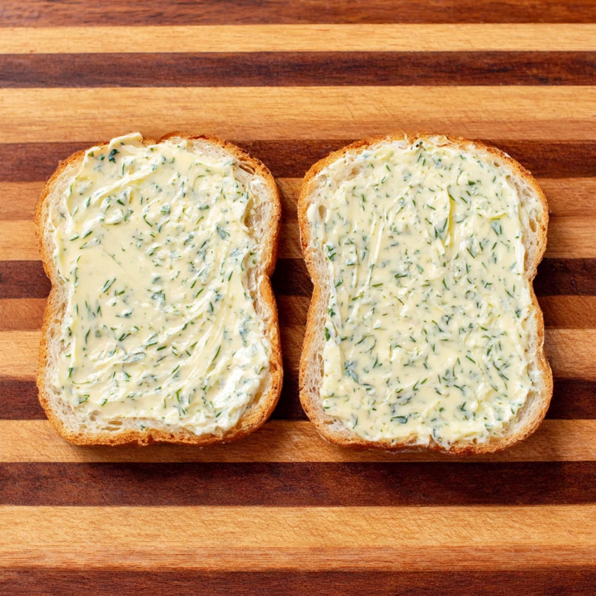 Two slices of bread spread evenly with creamy garlic and herb butter, ready for grilling on a wooden cutting board