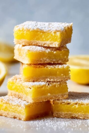 A close-up of stacked lemon bars, lightly dusted with powdered sugar, with fresh lemon slices in the background.