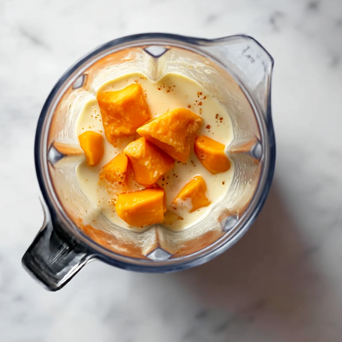 Overhead view of a blender with fresh mango chunks and creamy liquid, ready to be blended into a smooth and refreshing tropical mango smoothie.