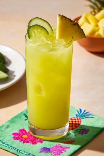 Tall glass of bright yellow-green Pineapple Cucumber juice with ice, garnished with pineapple wedge and cucumber slices, with fresh cucumber and diced pineapple in the background on a light table.