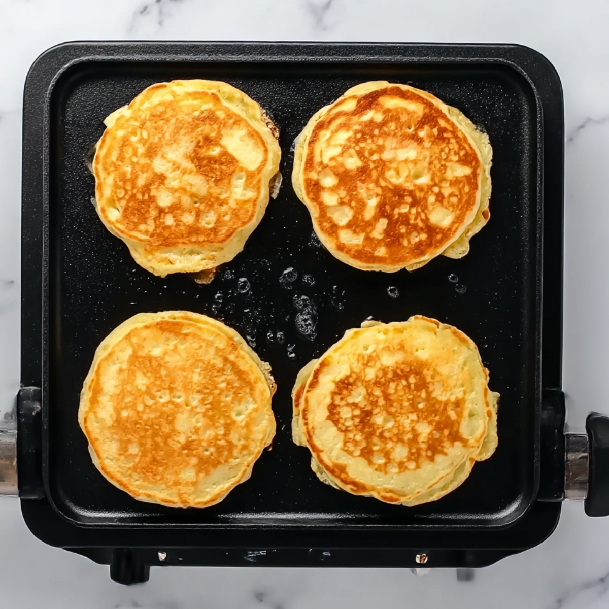 Four golden apple pancakes cooking on a black electric griddle, showing their perfectly browned surfaces and fluffy texture, ideal for a homemade breakfast recipe.