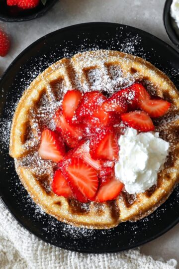 Close-up of a fluffy Belgian waffle topped with sliced fresh strawberries, whipped cream, and powdered sugar on a black plate, ready for a homemade breakfast or brunch.