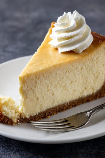 A slice of rich and creamy cheesecake with a golden graham cracker crust, topped with a swirl of whipped cream, served on a white plate with a fork.