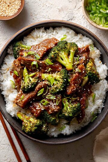 A bowl of tender beef and vibrant broccoli stir-fry served over fluffy white rice, garnished with sesame seeds and sliced green onions, with small bowls of sesame seeds and chopped green onions on the side.
