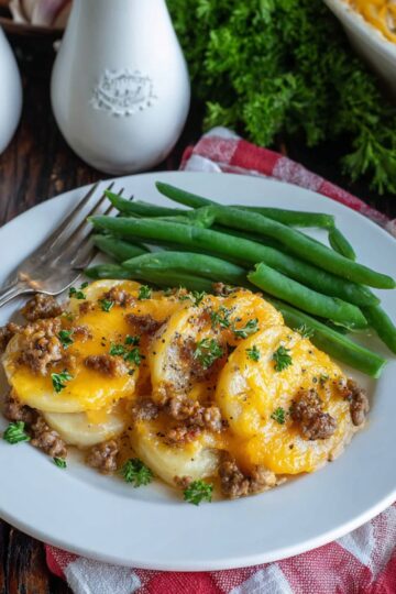 A plate of cheesy hamburger potato casserole topped with melted cheddar and parsley, served with fresh steamed green beans on a white plate with a red checkered napkin.