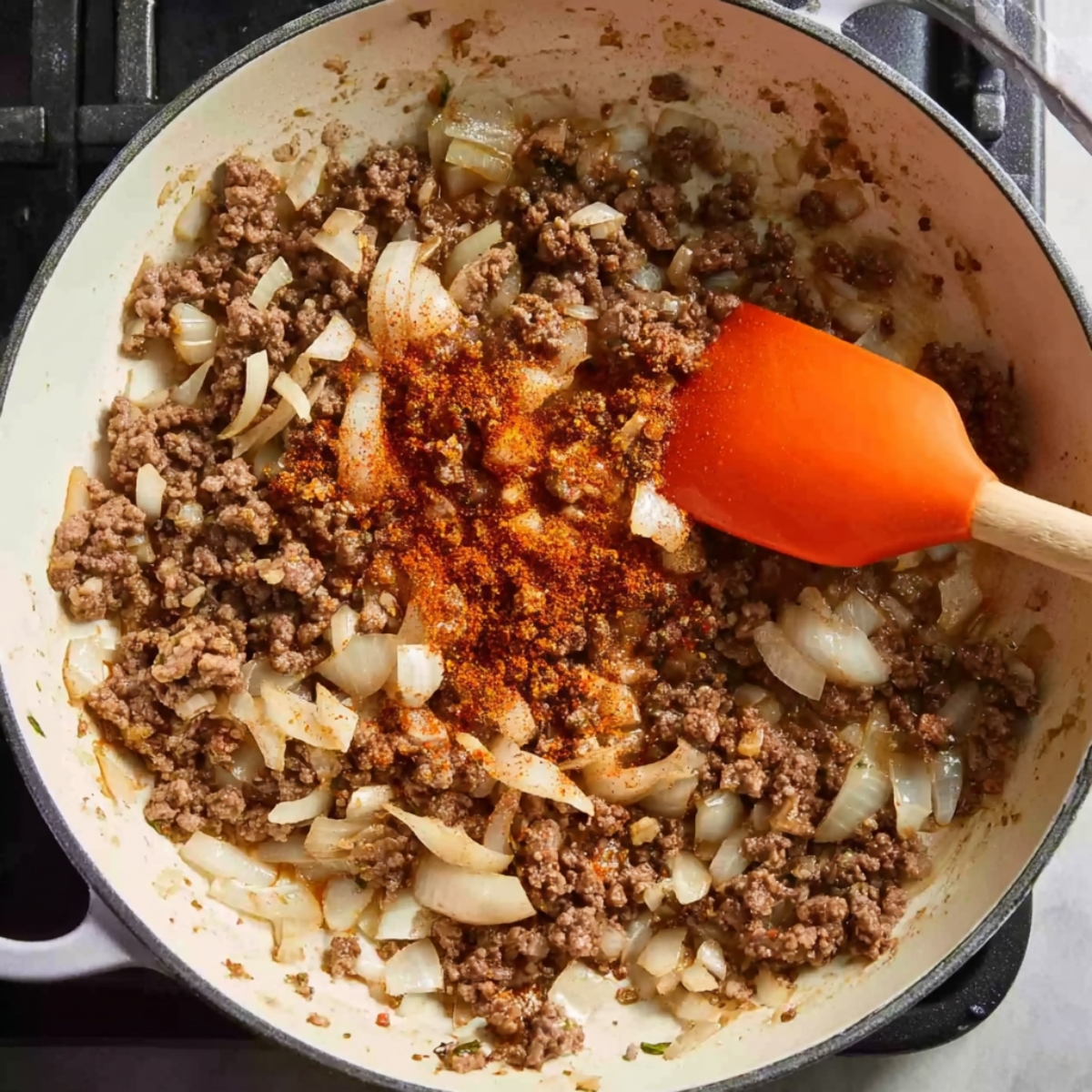 Ground beef and chopped onions cooking in a skillet with paprika and spices, being stirred with an orange spatula for a cheesy hamburger potato casserole.