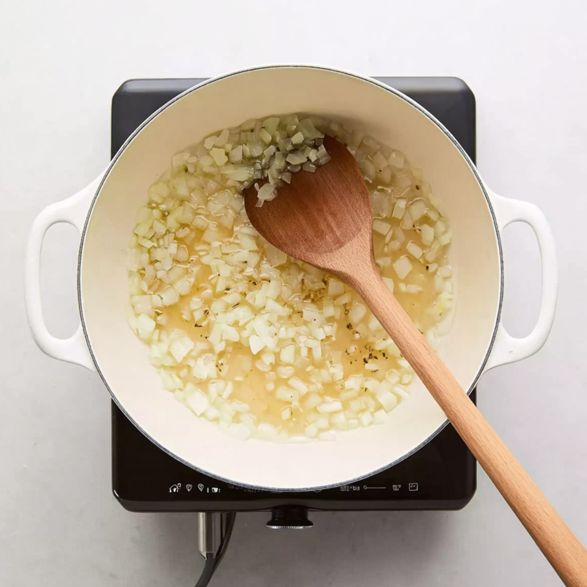 Chopped onions being sautéed in a white pot with oil and seasonings on a stovetop, stirred with a wooden spoon, for a classic goulash recipe.