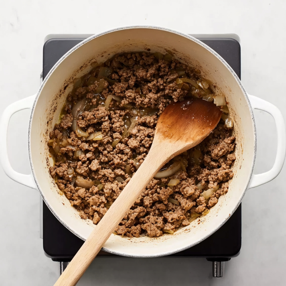Ground beef cooking with onions in a white pot on a stovetop, stirred with a wooden spoon, for a classic goulash recipe.