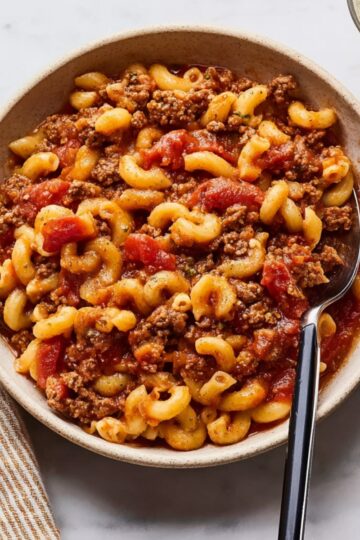 Bowl of classic goulash with tender ground beef, elbow macaroni, and rich tomato sauce, garnished with herbs, served with a side drink on a light background.