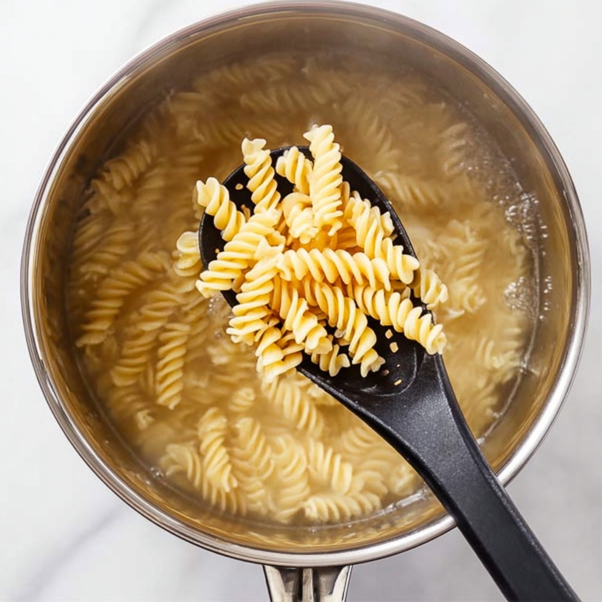 Top-down view of rotini pasta being boiled in a stainless steel pot, held on a black slotted spoon, as the first step for preparing a creamy chicken casserole.