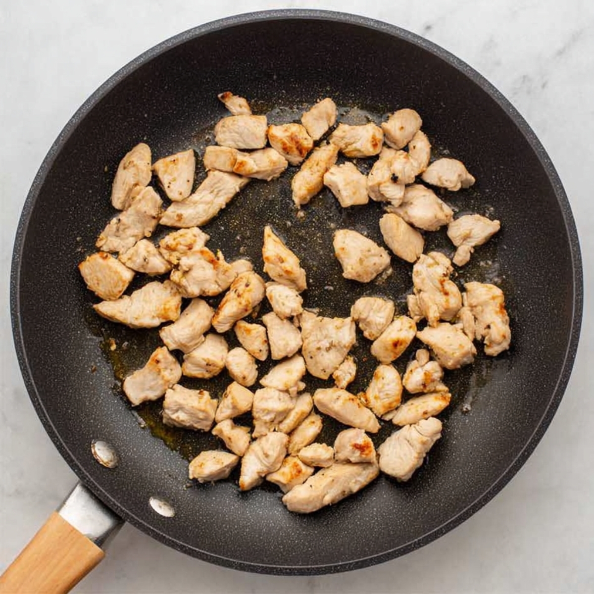 Top-down view of bite-sized chicken pieces being sautéed in a black nonstick skillet, lightly browned and cooked, ready to be added to a creamy chicken casserole.