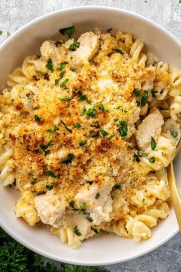 Close-up of a creamy chicken casserole made with rotini pasta, tender chicken chunks, a cheesy sauce, and a crispy golden breadcrumb topping, garnished with fresh parsley in a white bowl.