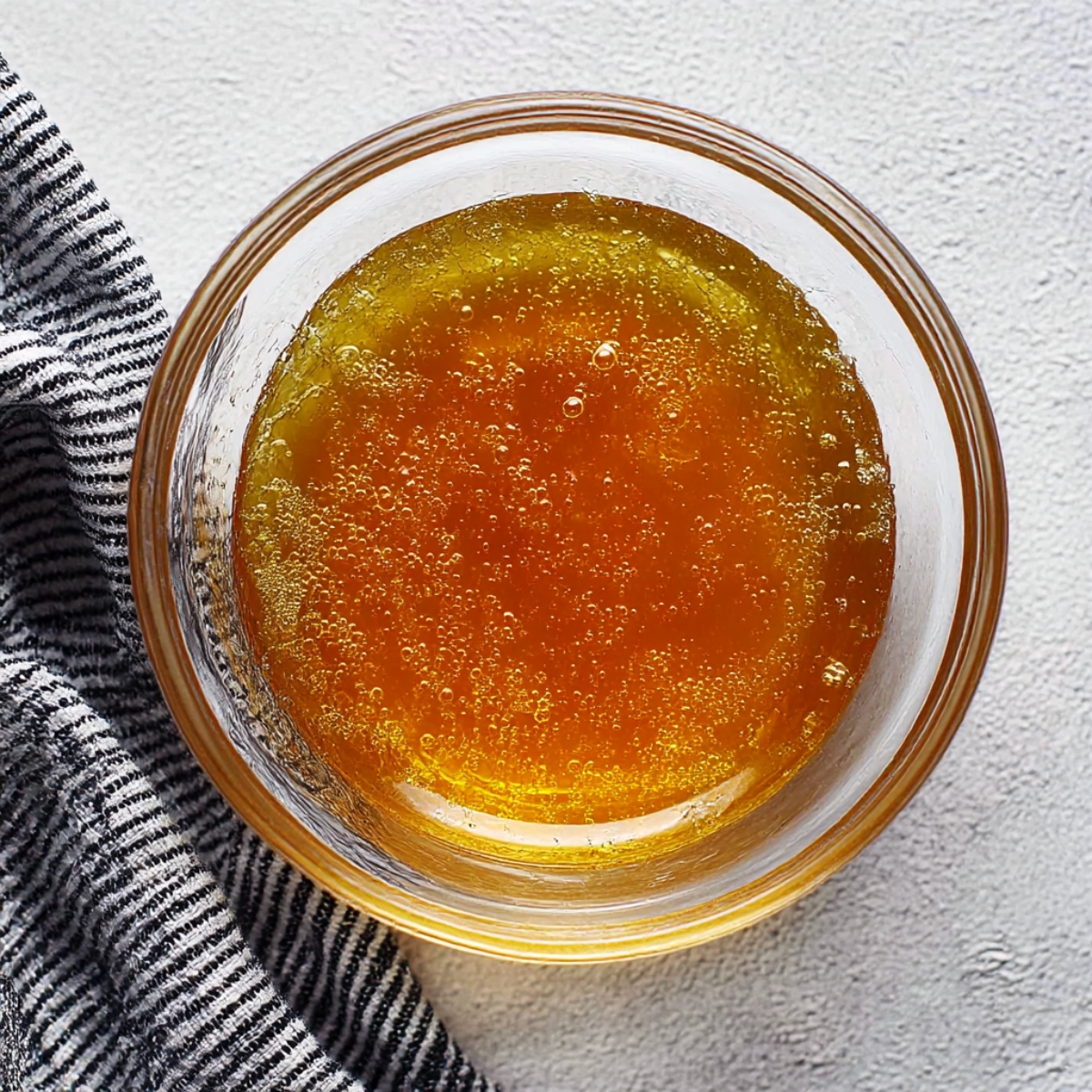 Top-down view of a small glass bowl filled with golden coconut syrup, next to a striped cloth on a light surface, ready to drizzle over creamy coconut rice pudding.