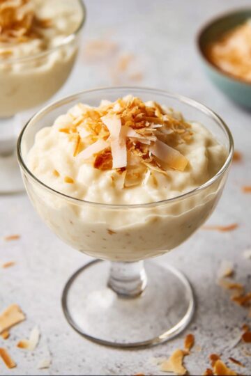 A glass dessert cup filled with creamy coconut rice pudding, topped with toasted coconut flakes, on a light surface with scattered coconut around.