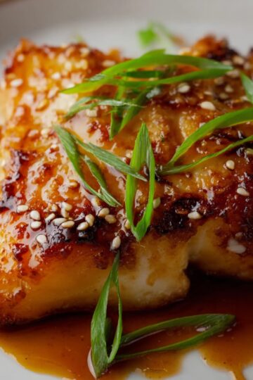 Close-up of a golden-brown honey-soy glazed cod fillet, garnished with sesame seeds and fresh green onions, served on a white plate with rich savory glaze.