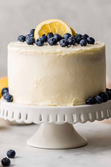 A tall lemon blueberry layer cake frosted with creamy cream cheese frosting, topped with fresh blueberries and lemon slices, displayed on a white cake stand.