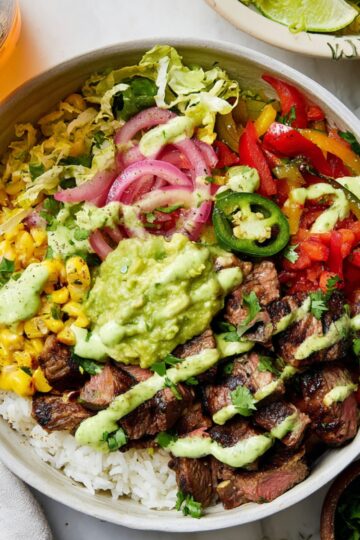 Overhead view of a vibrant Mexicali Steak Burrito Bowl with tender grilled steak slices, white rice, charred corn, colorful bell peppers, pickled red onions, fresh shredded lettuce, jalapeño slices, creamy guacamole, and drizzled cilantro lime dressing in a bowl, perfect for a quick Mexican-inspired meal.