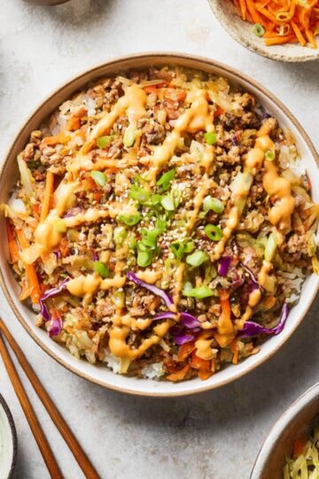 A bowl of homemade Egg Roll in a Bowl with ground beef, shredded cabbage, carrots, and green onions, drizzled with a creamy sesame sauce, served with chopsticks on the side.