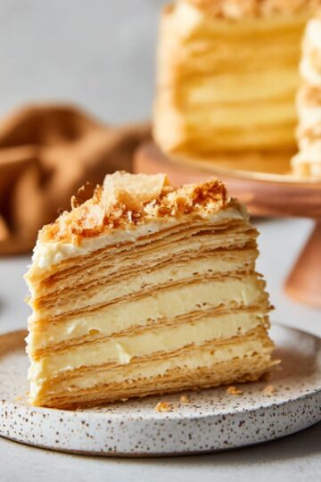 A slice of classic Napoleon Cake showing multiple flaky puff pastry layers filled with creamy custard, served on a speckled ceramic plate with crumbs on top, perfect for a homemade dessert.