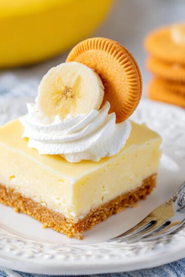 Delicious no-bake banana cheesecake slice topped with whipped cream, a banana slice, and a golden cookie on a white plate, perfect for easy homemade dessert recipes.