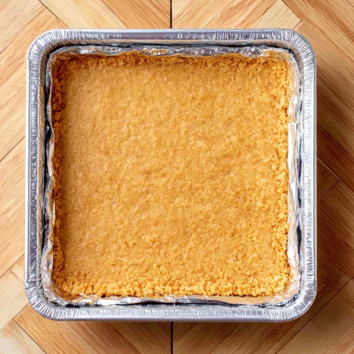 Close-up of a perfectly pressed golden graham cracker crust in a square pan, ready for a no-bake banana cheesecake filling.