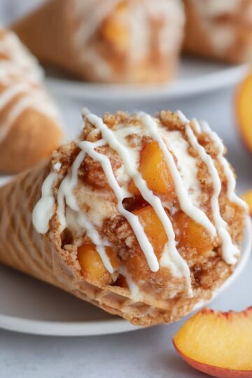 Close-up of a peach cobbler cheesecake cone with chunks of fresh peaches, creamy cheesecake filling, cinnamon crumble, and a drizzle of white icing, served on a plate with sliced peaches.